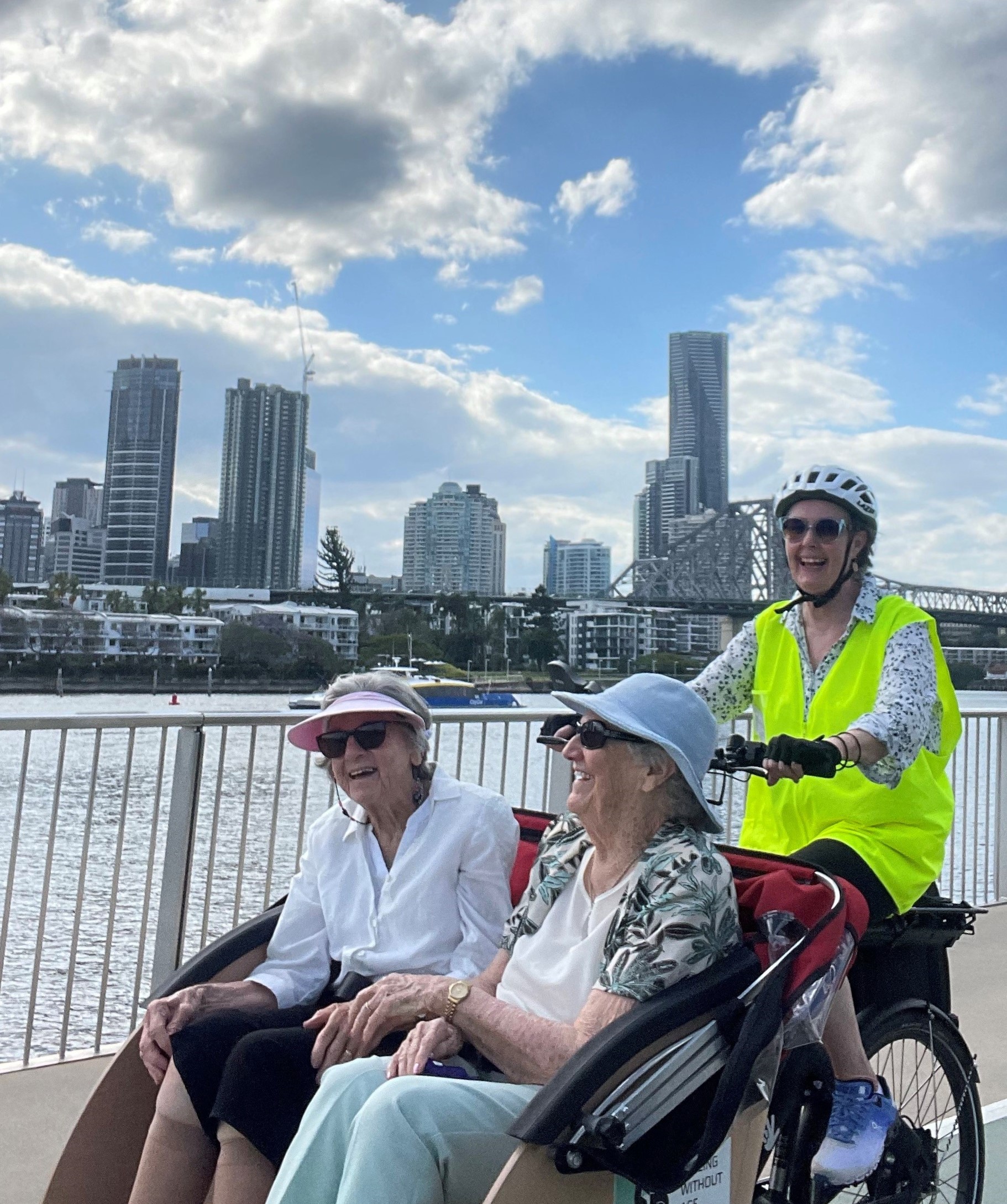 two ladies sitting in a carriage on the front of a bike with a lady pedaling behind them across a bridge